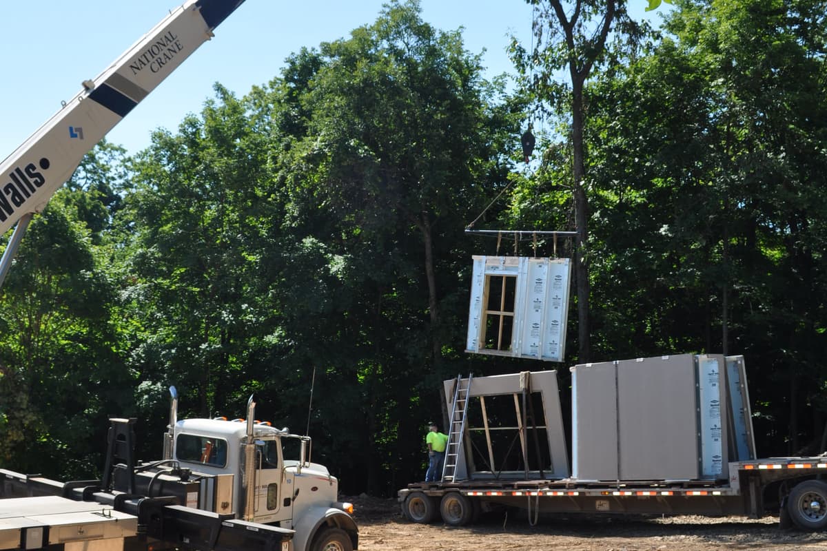 A panel swinging on the crane hook over the pad, the boom truck braced below and the woods behind.