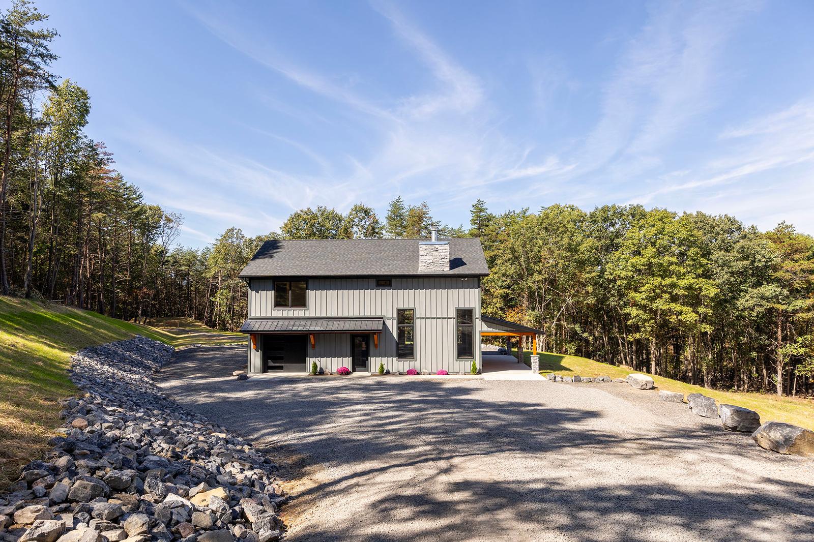 Two-story modern farmhouse in gray board-and-batten with a stone chimney and covered patio, above a wide gravel drive