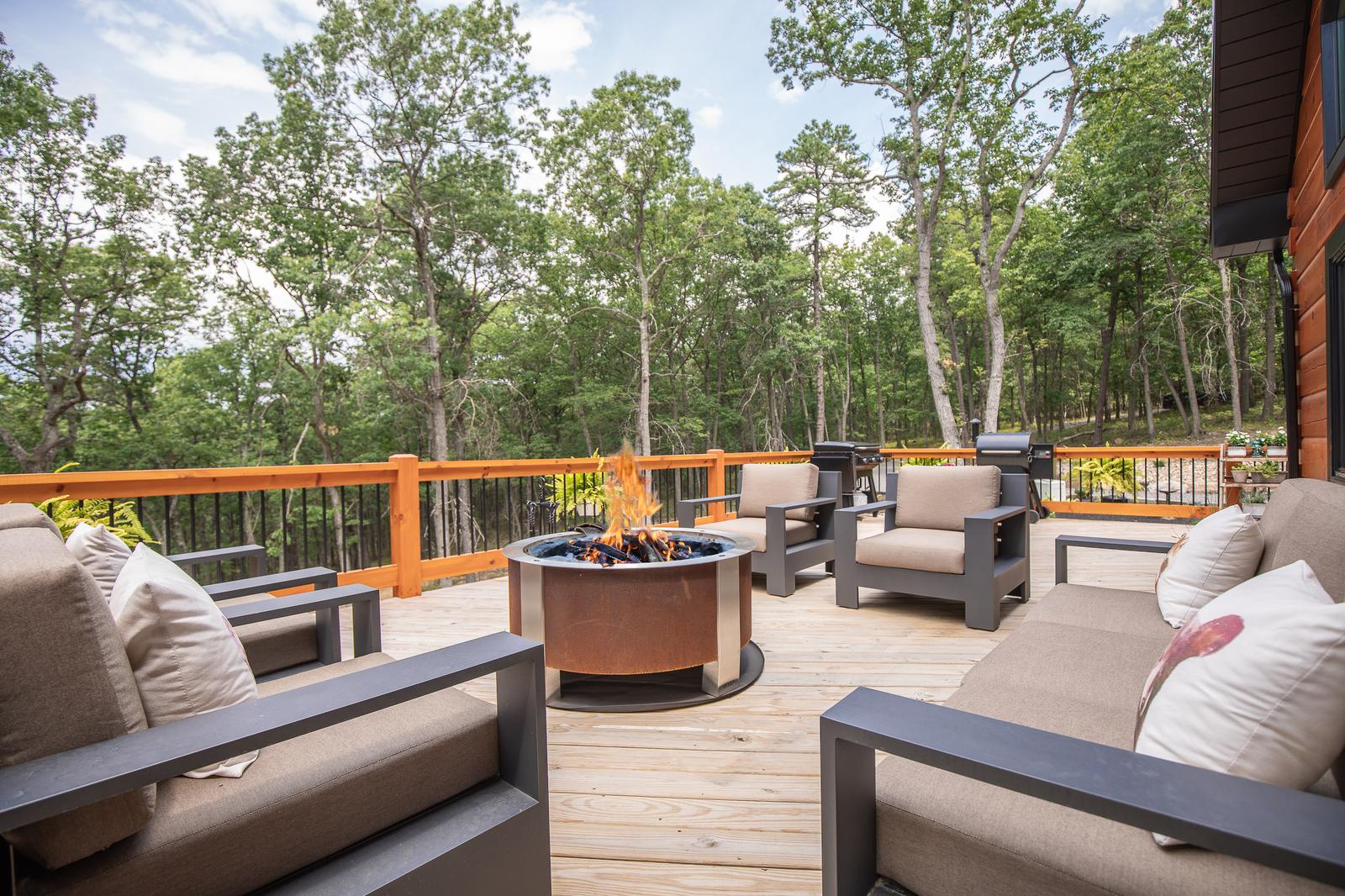Deck with a lit fire pit, cushioned seating and stained wood railing, with forest beyond in daylight
