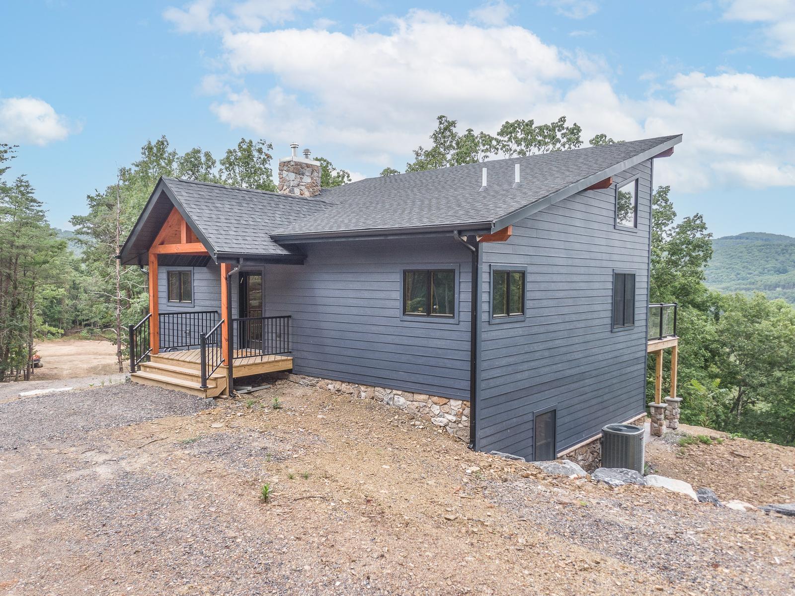 Front exterior of a hybrid home with gray siding, timber entry accents, and a wooded ridge behind