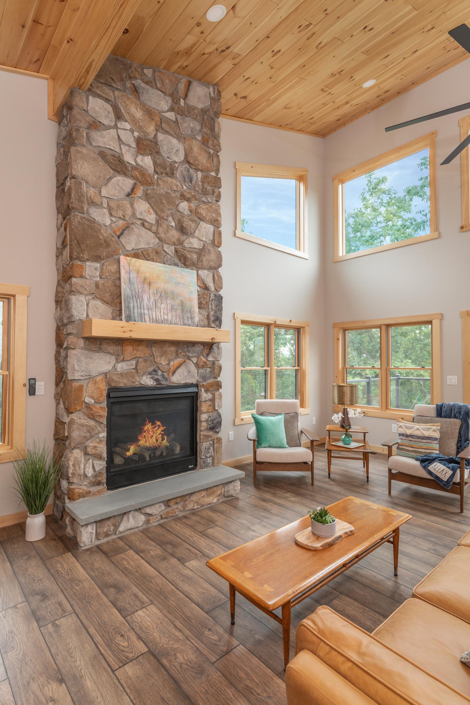 Tall stone fireplace with a lit fire, timber mantel and painting, beneath a vaulted pine ceiling
