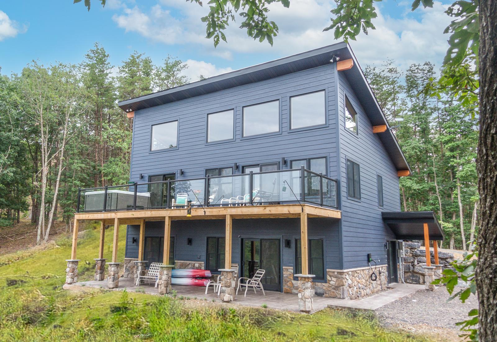 Rear exterior showing an elevated deck on stone piers with glass railing and a hot tub, surrounded by forest