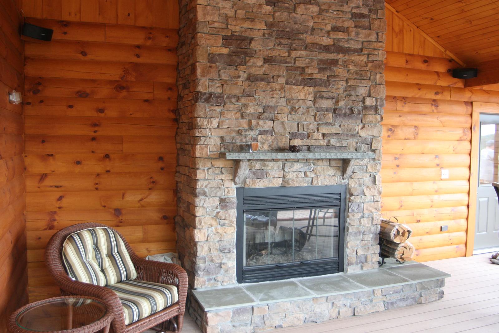 An outdoor stone fireplace on a covered porch, a striped wicker armchair beside it and split logs stacked on the slate hearth
