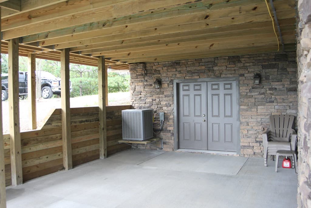 The area under the deck, floored in concrete between stone piers, with a pair of gray doors into the lower level and the air-conditioning condenser on a bracket beside them