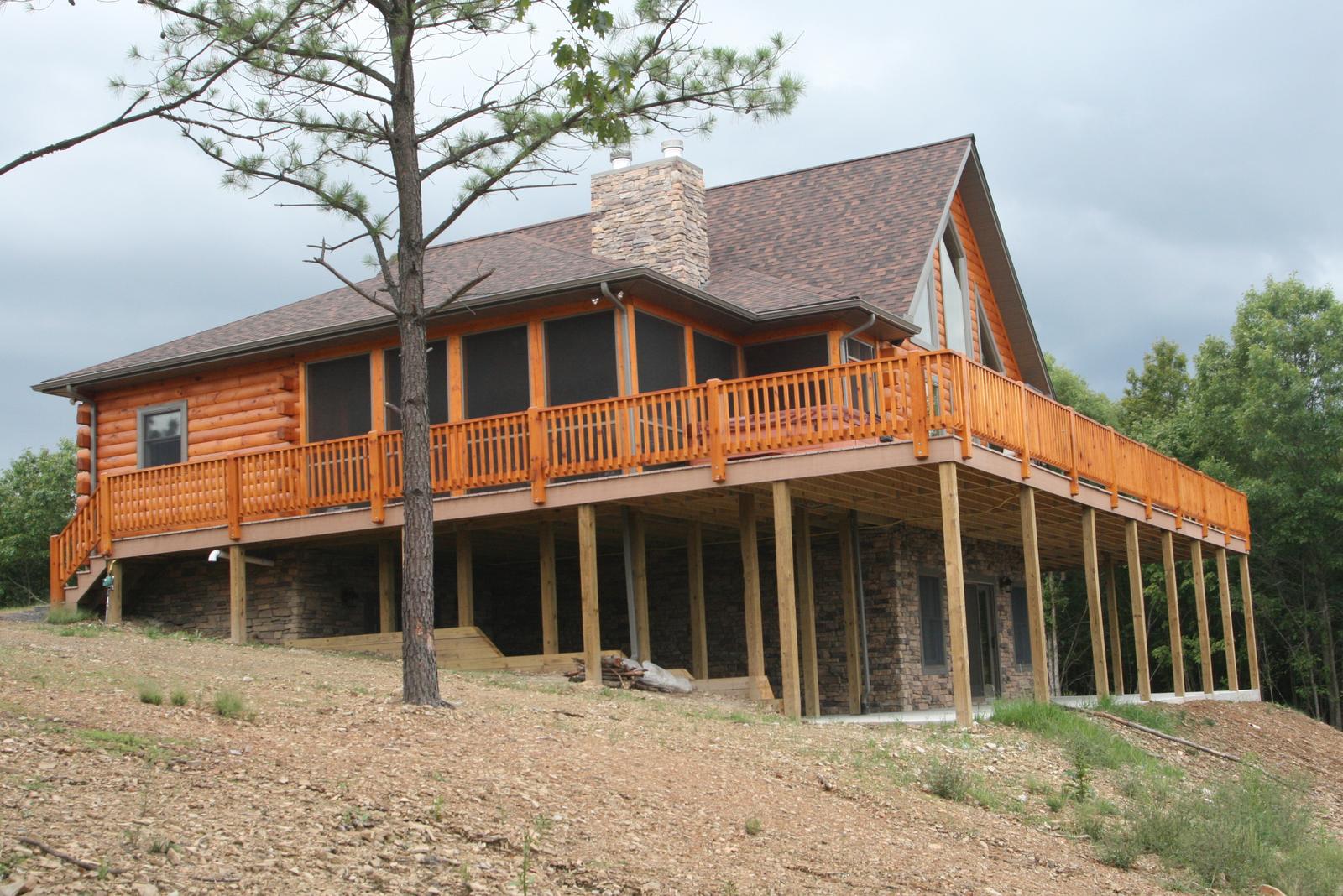 A log home on a bare graded slope, a stone chimney rising the full height of the near gable and a wide deck with a log railing carried on posts above a stone-faced lower level