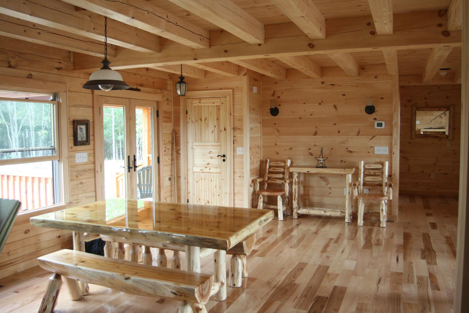 A dining area in log walls with a peeled-log table and bench on hardwood floors, pendant lamps under exposed joists and glazed doors to the deck
