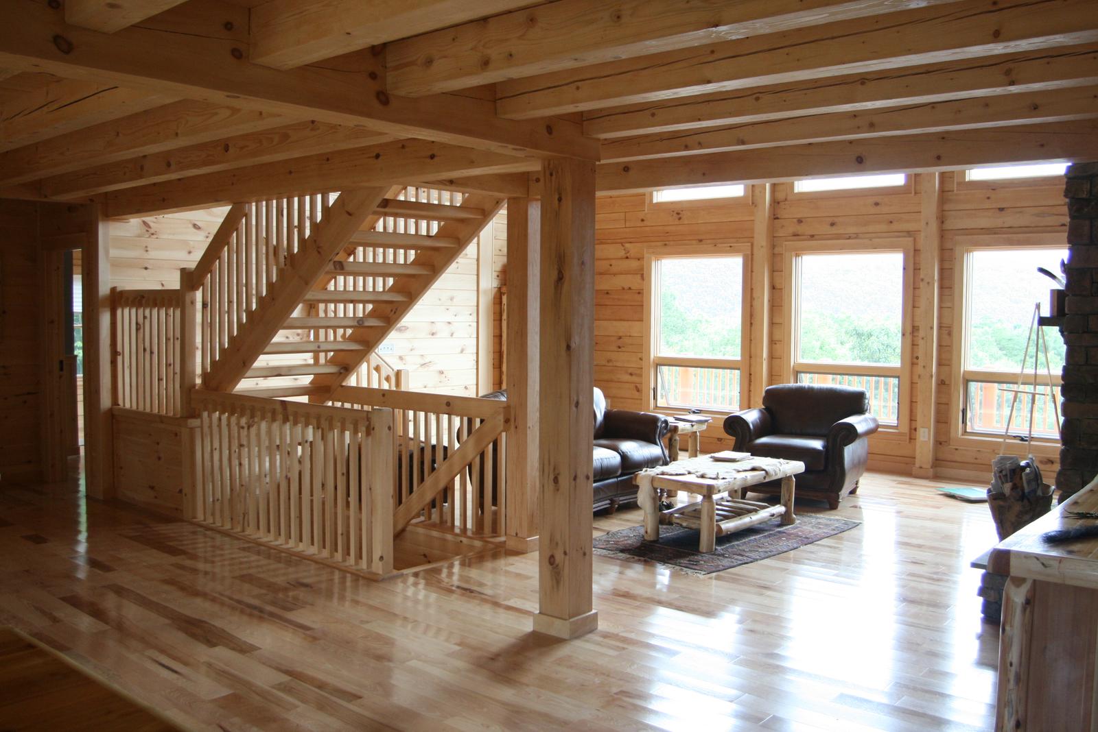 An open stair with slender square balusters seen across a great room, leather chairs at a wall of windows and the stone chimney at the right edge