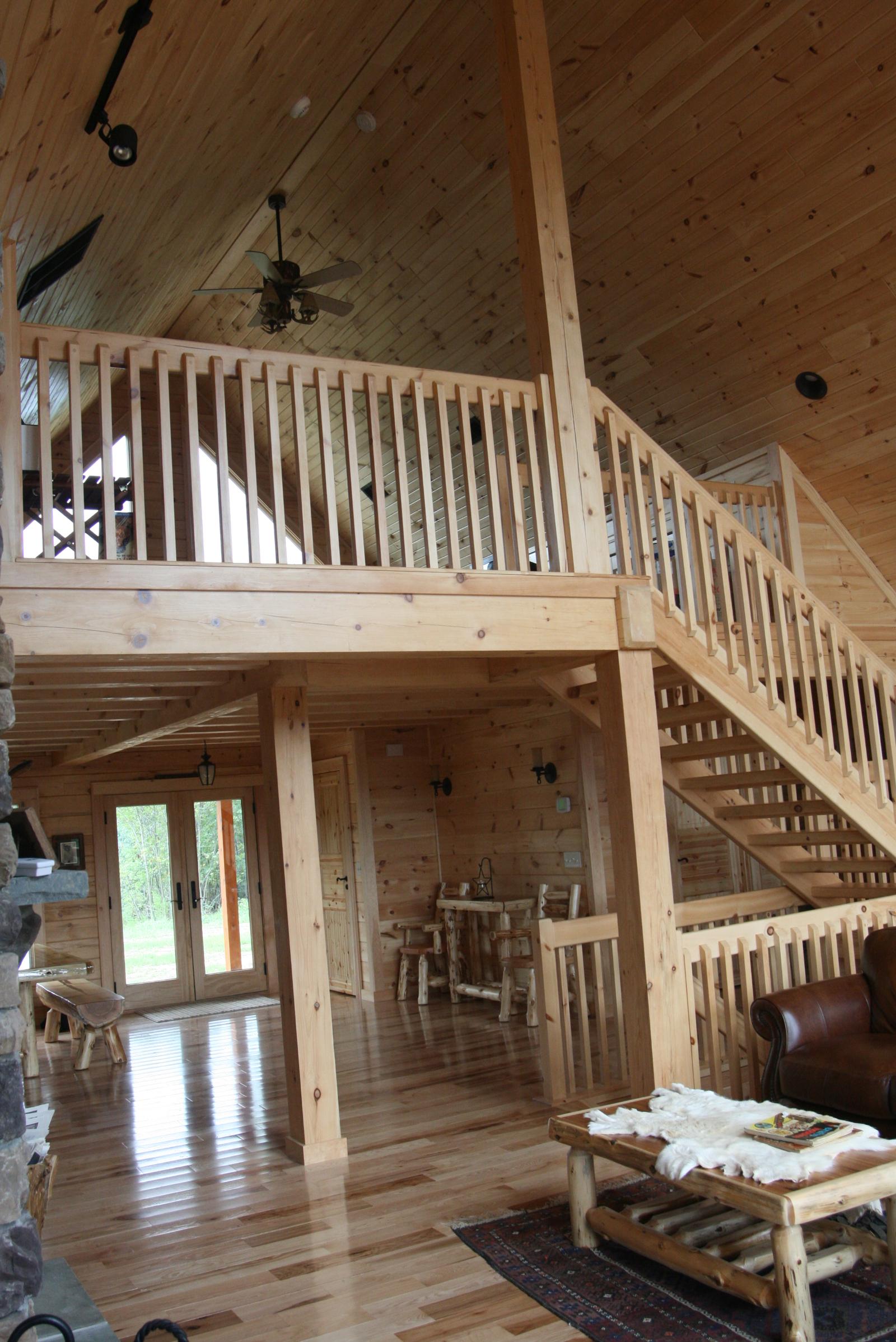 A loft balcony carried on square posts above an open floor, the stair rising at the right and glazed doors to the lawn at the far end