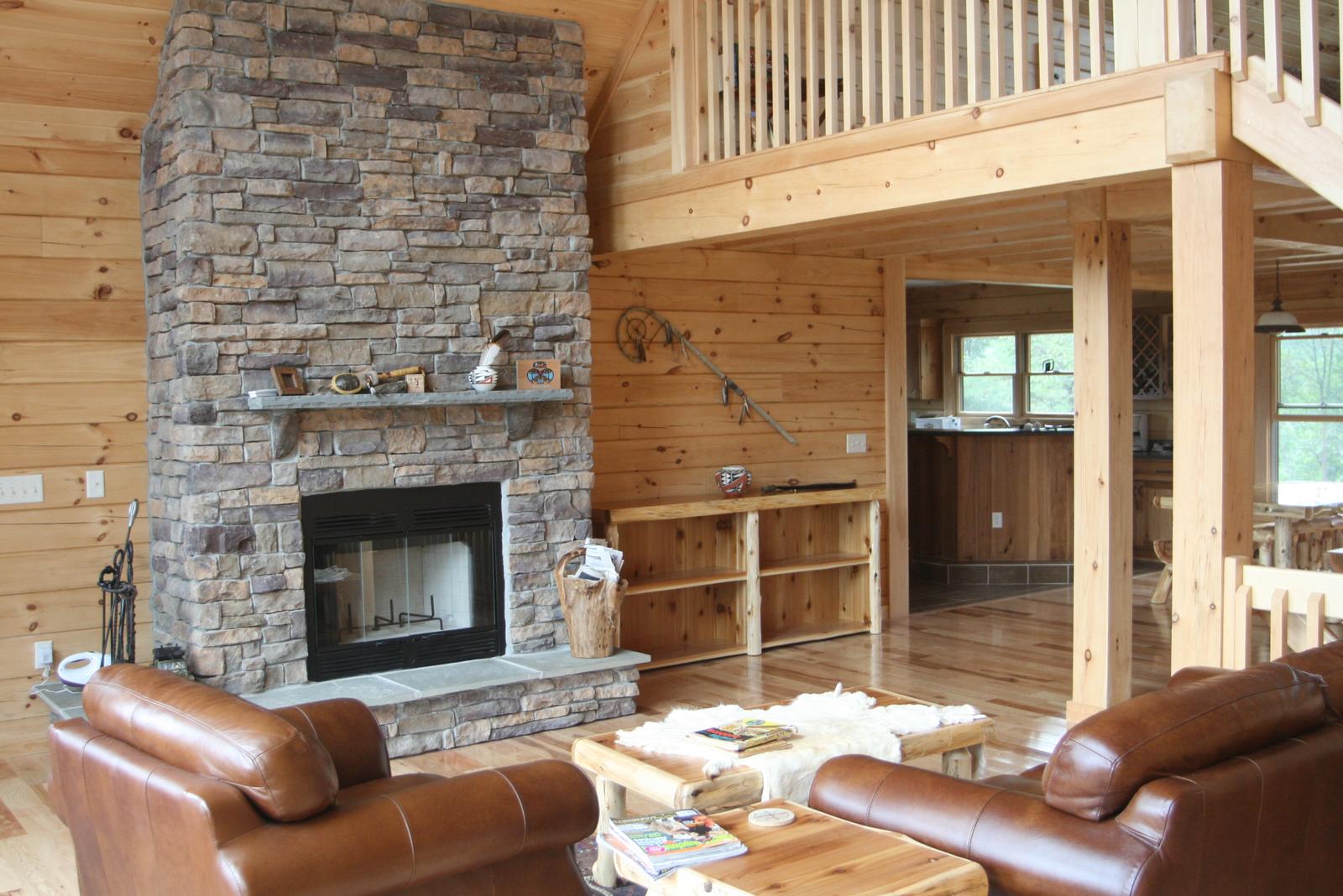A stone fireplace running the full height of the great room to the loft rail, leather sofas around a log table and the kitchen through the opening beyond