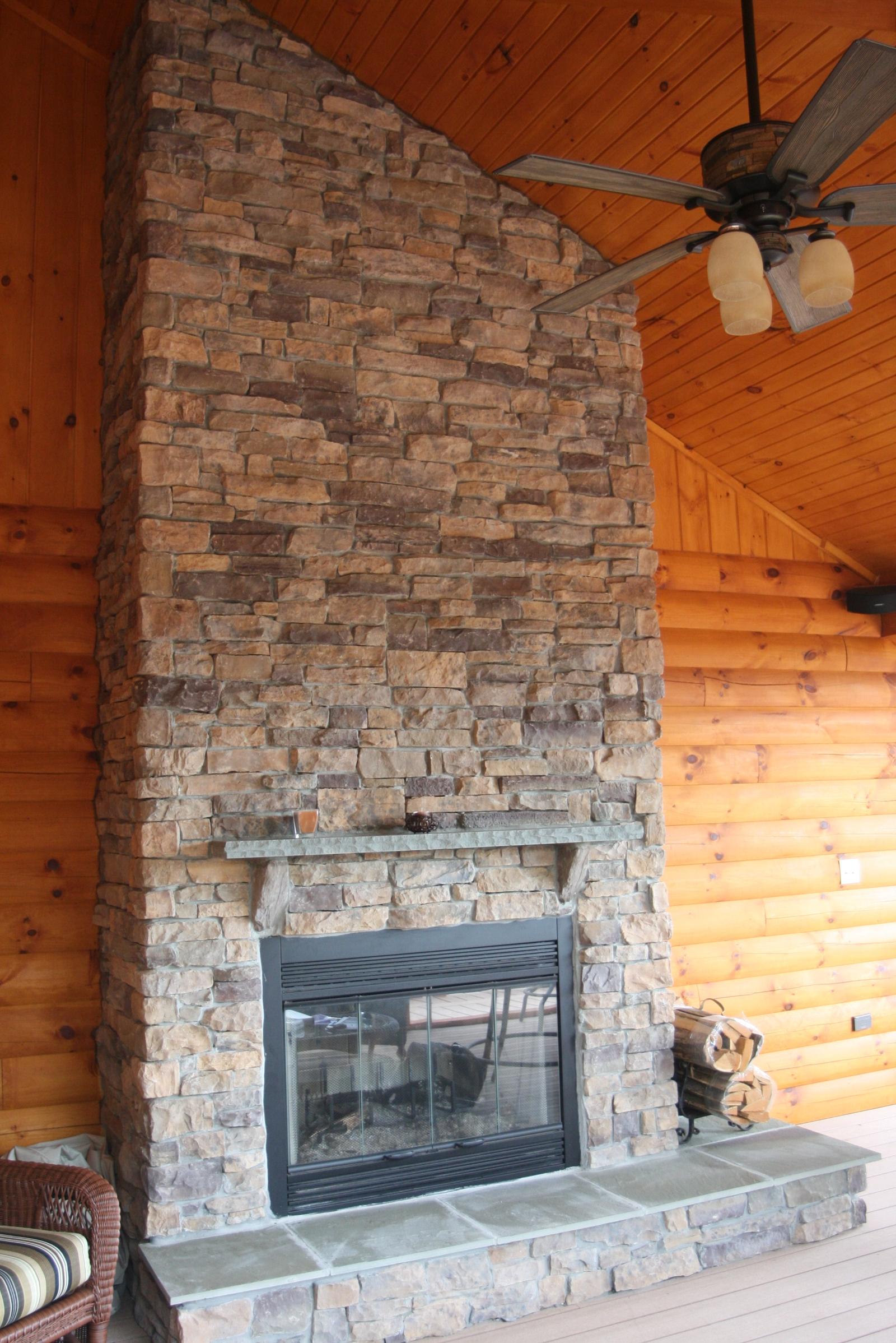 A stone chimney rising to a vaulted board ceiling on a covered porch, a black fireplace insert below a slate mantel and a ceiling fan overhead