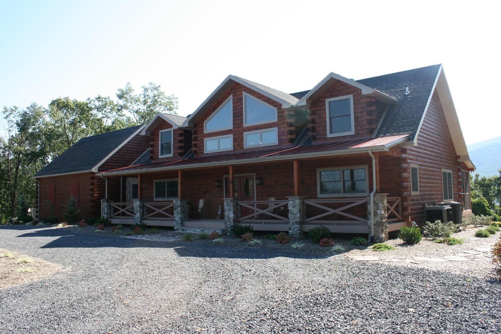 A log home with two roof dormers and a covered front porch on stone piers, its railing built in a repeated X pattern, a gravel drive running up to it
