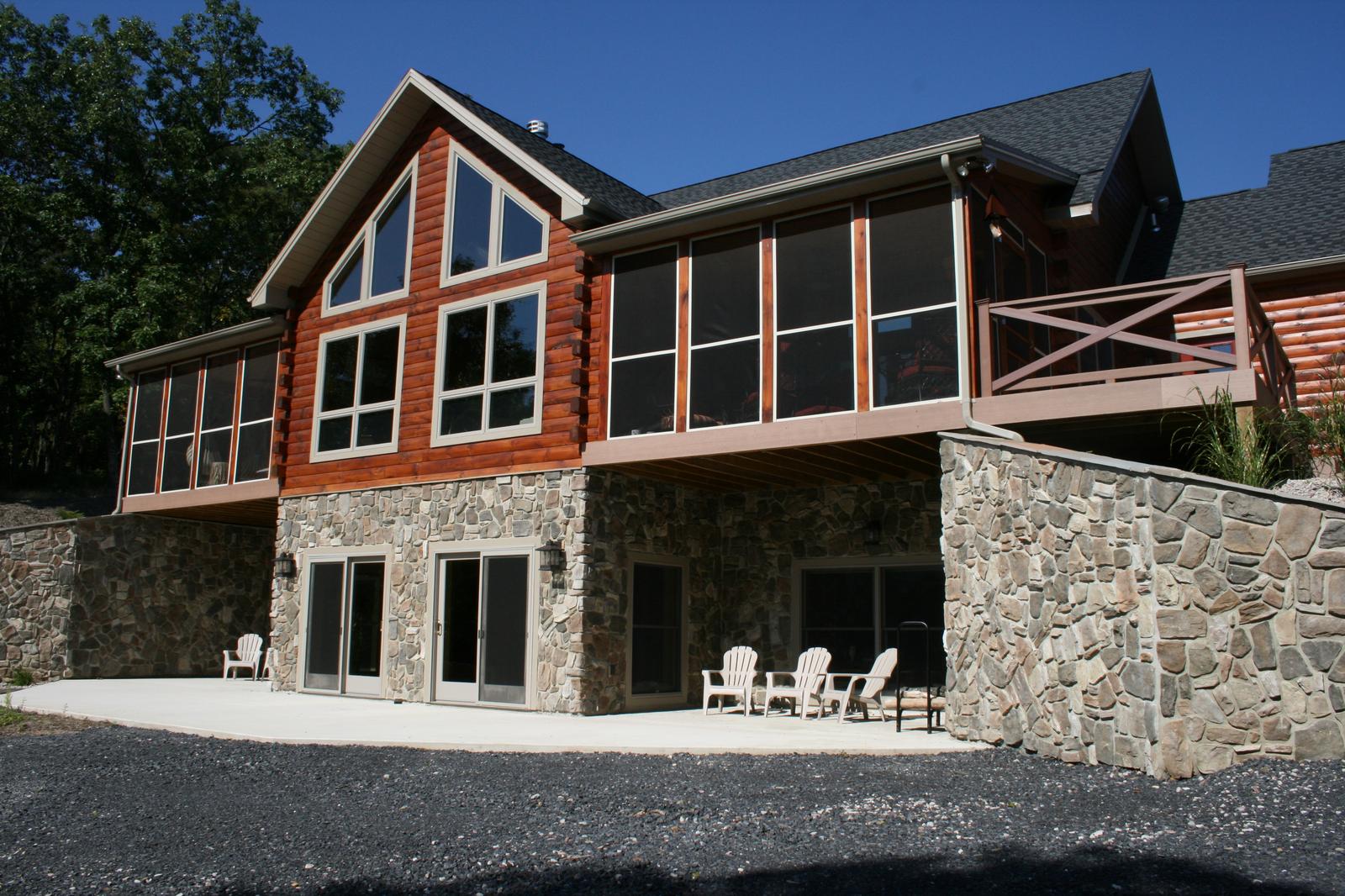The back of a log home with a stone walkout below a screened porch, a prow gable of windows above it and chairs out on the patio