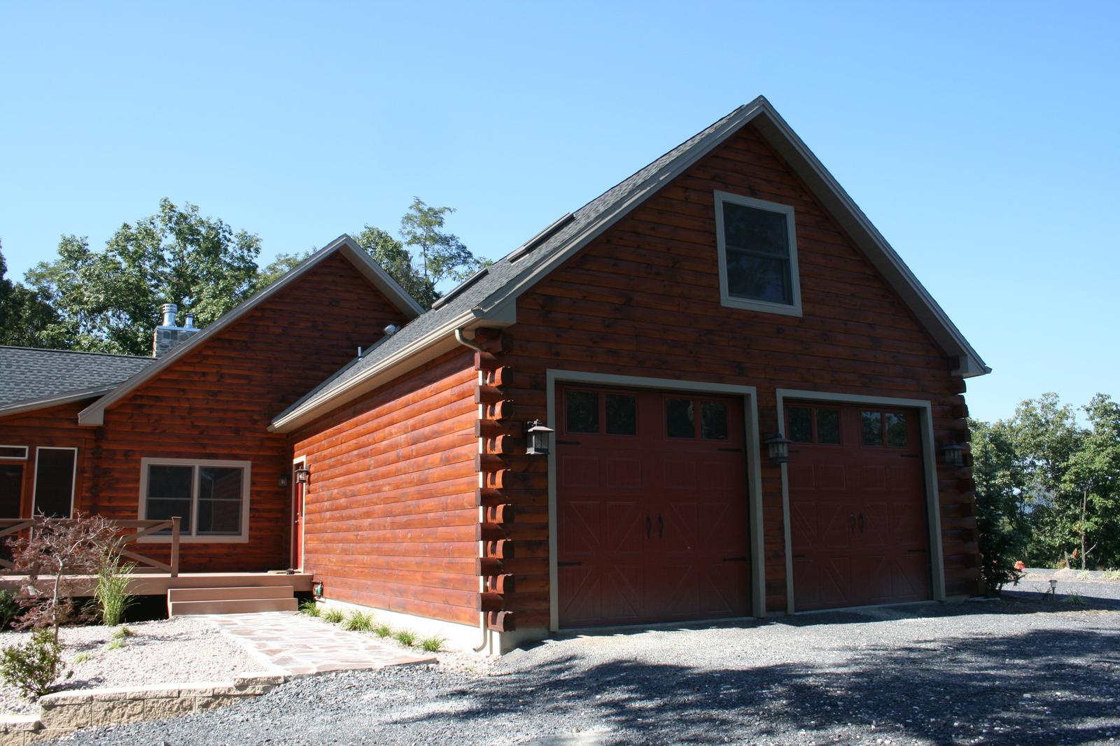 A log garage with two red doors and a gable window, the main house stepping down to the left across a gravel apron