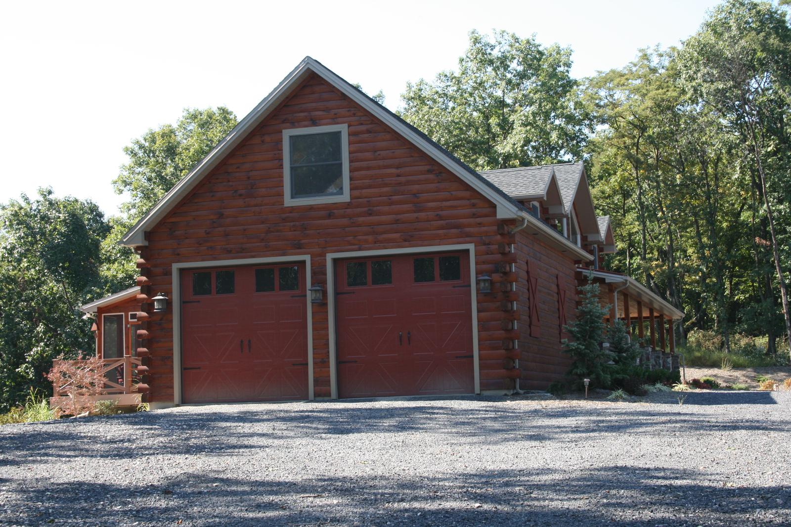 A red-stained log house with two carriage-style garage doors under a gable, dormers running away to the right and a gravel drive across the front