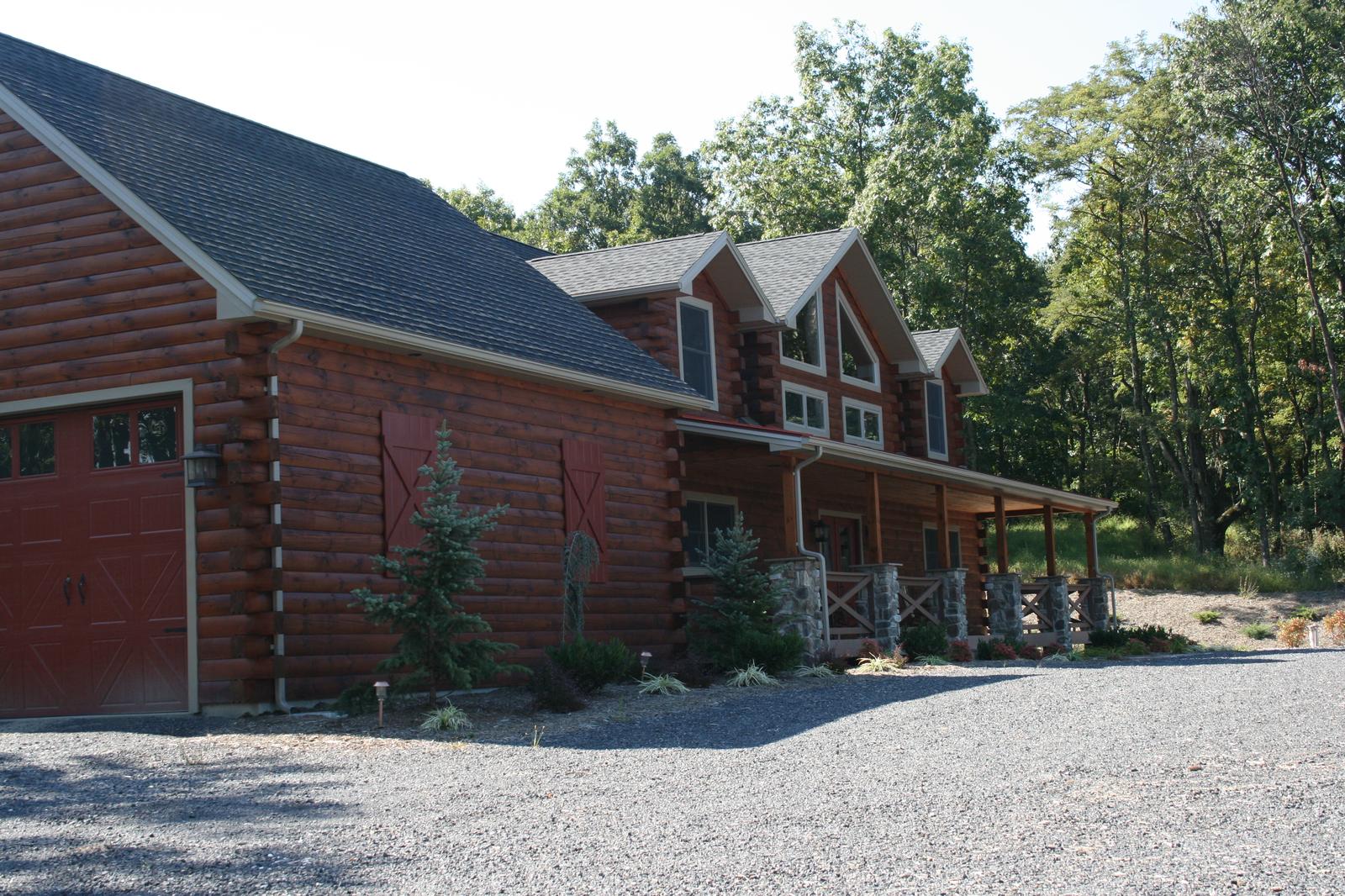 The long side of a log home seen from the garage end, three dormers above a covered porch carried on stone piers