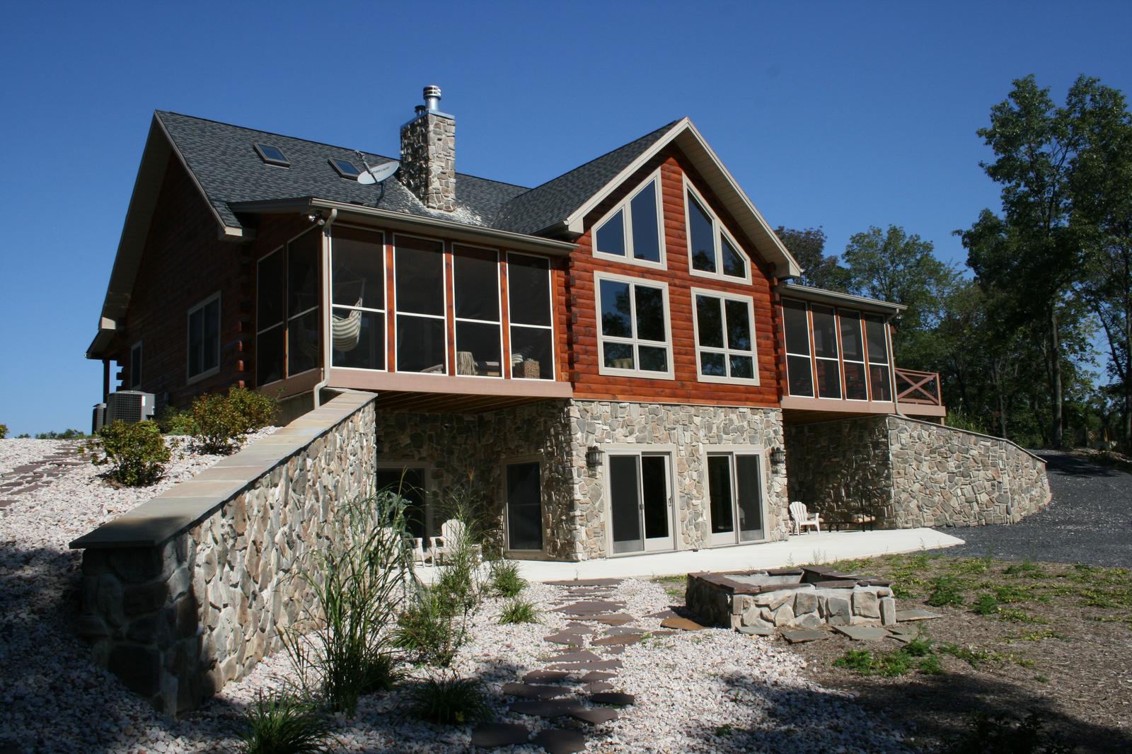 A house with a screened porch under a gable of glass and timber, a stone lower level opening onto a patio, with stone retaining walls stepping down through the landscaping