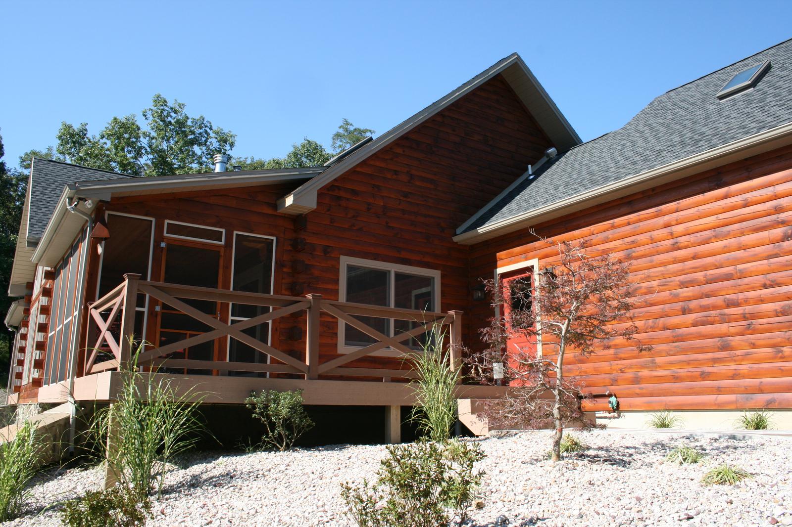 A screened porch with an X-braced rail at the corner of a log home, a red door beside it and white gravel beds below