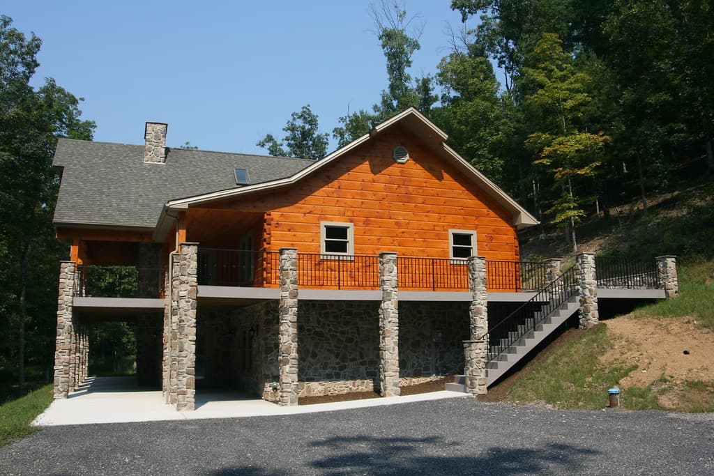 The gable end from the drive, log wall above a run of stone piers, a deck and open stair coming down to gravel