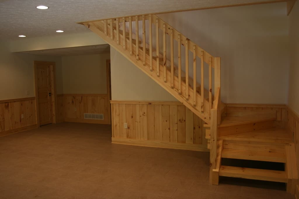 A finished lower-level room, the stair coming down along one wall above board wainscot, floor bare and ready