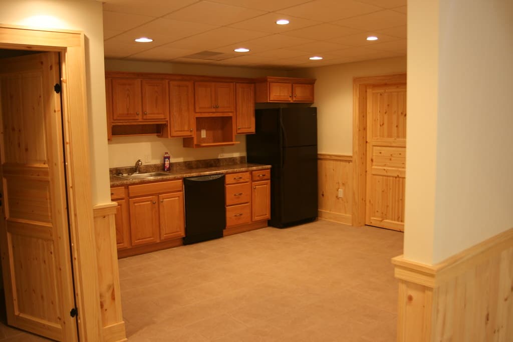 A lower-level kitchenette from the doorway, wall cabinets and dark appliances against board wainscot
