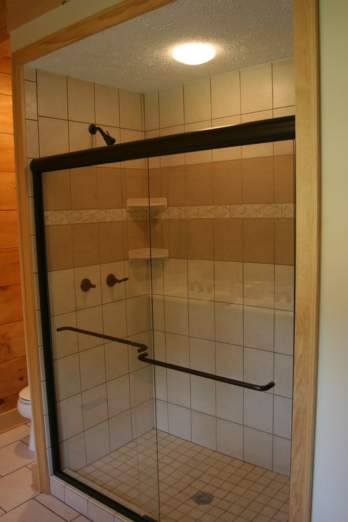 A tiled shower with a framed glass door, corner shelves and a long grab bar, set into a boarded wall