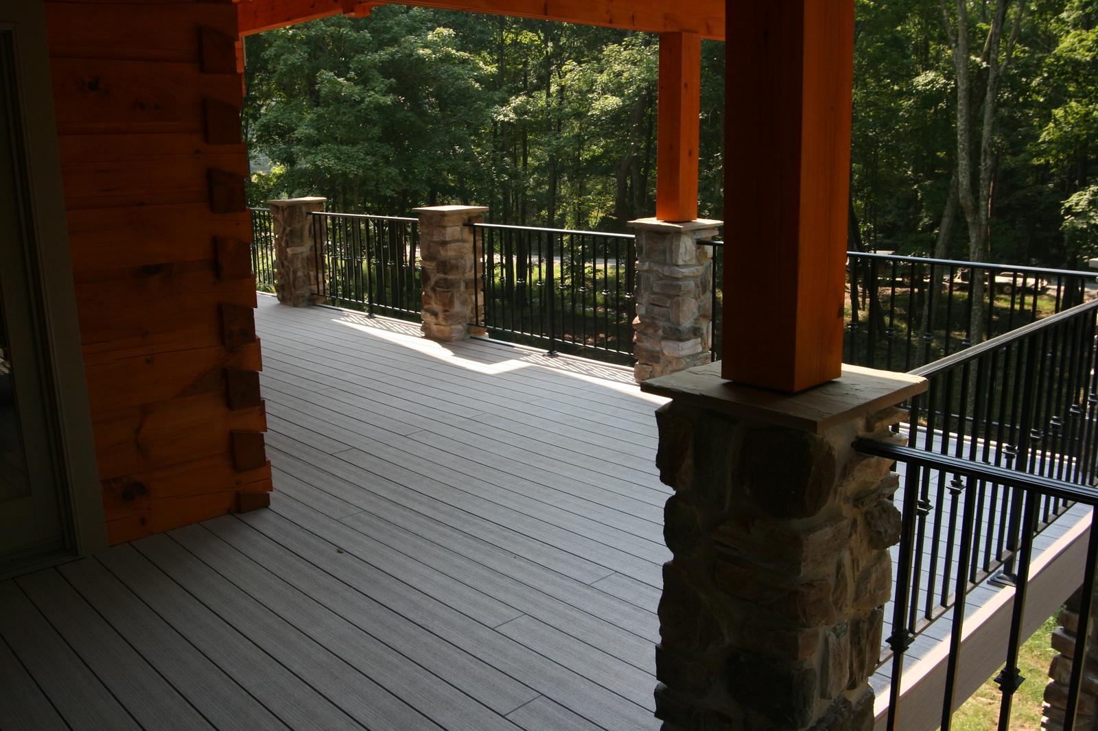 A covered porch running past stone piers and a black metal railing, green woods filling the opening