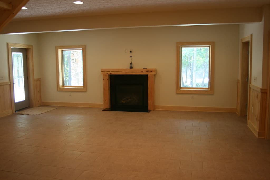 A lower-level room with a fireplace and plain mantel between two windows, tile floor, nothing in it yet