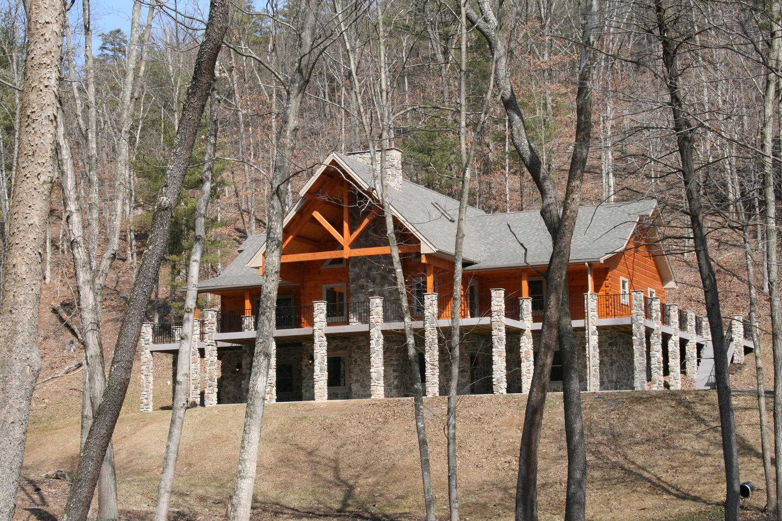A log home in bare woods, a heavy timber truss filling the entry gable above a covered porch on stone piers