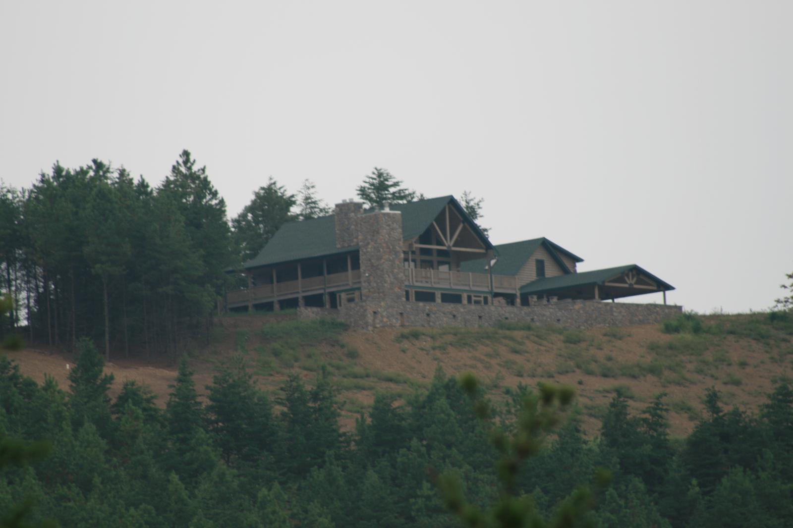 A long house on a ridge seen from far below across a wooded hillside, its green metal roof and stone chimney above a covered porch, with a stone wall running the length of the front