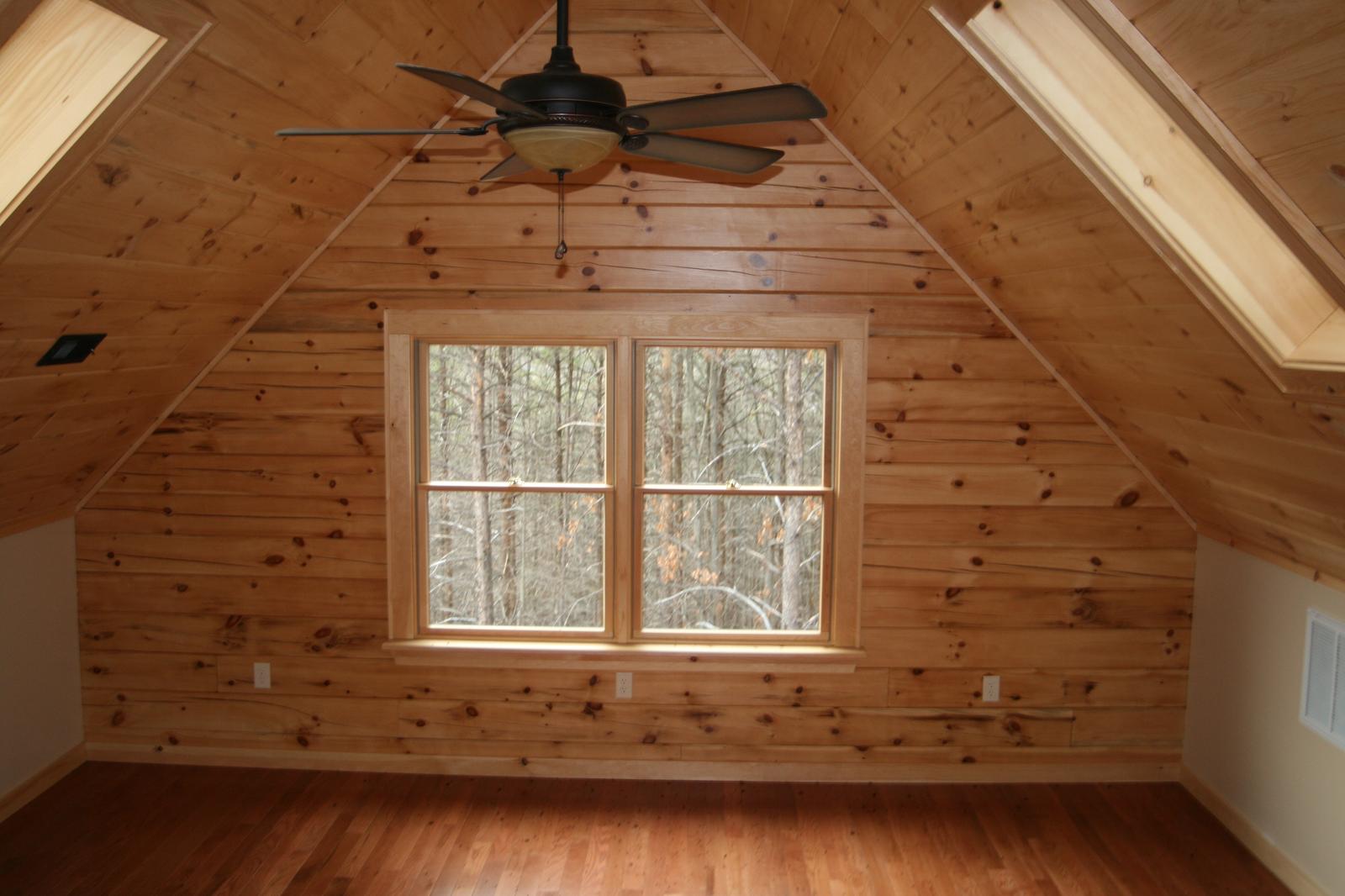 A loft bedroom under a sloped board ceiling, a fan at the ridge and a window in the gable