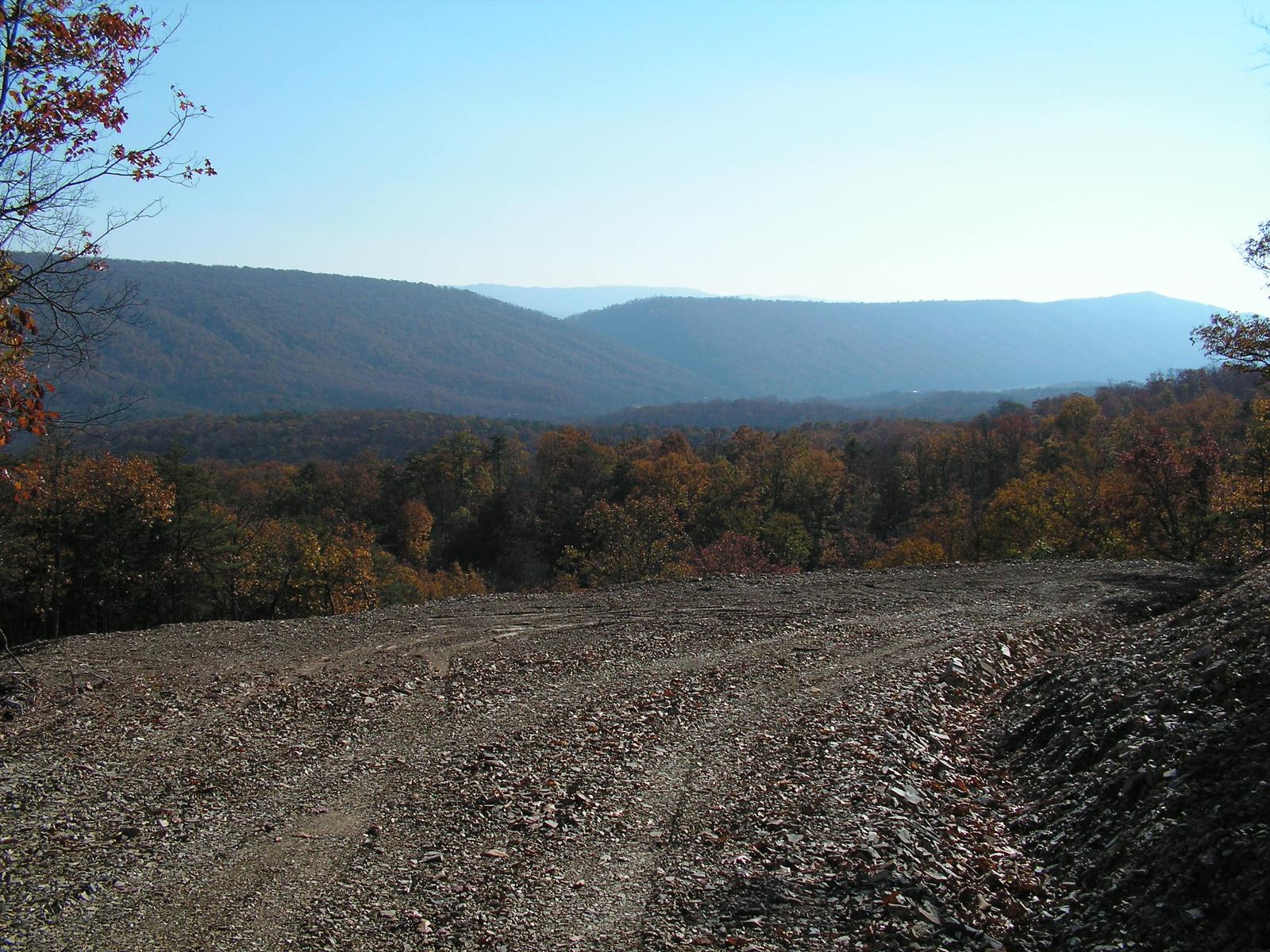 A wide graded pad on a ridge top, machine tracks curving across the fresh surface, layered blue mountains beyond the fall treetops