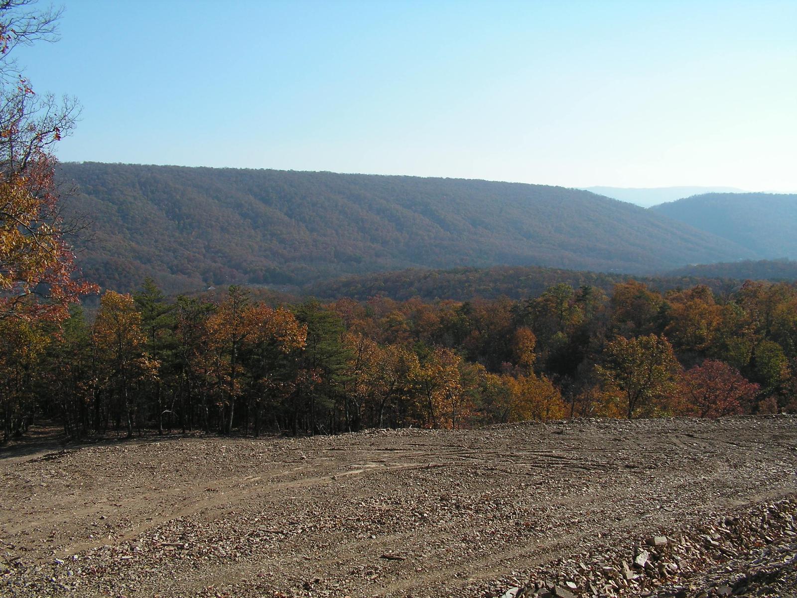 A broad level building pad cut into a ridge, its stone edge in the foreground and a long forested mountain across the valley
