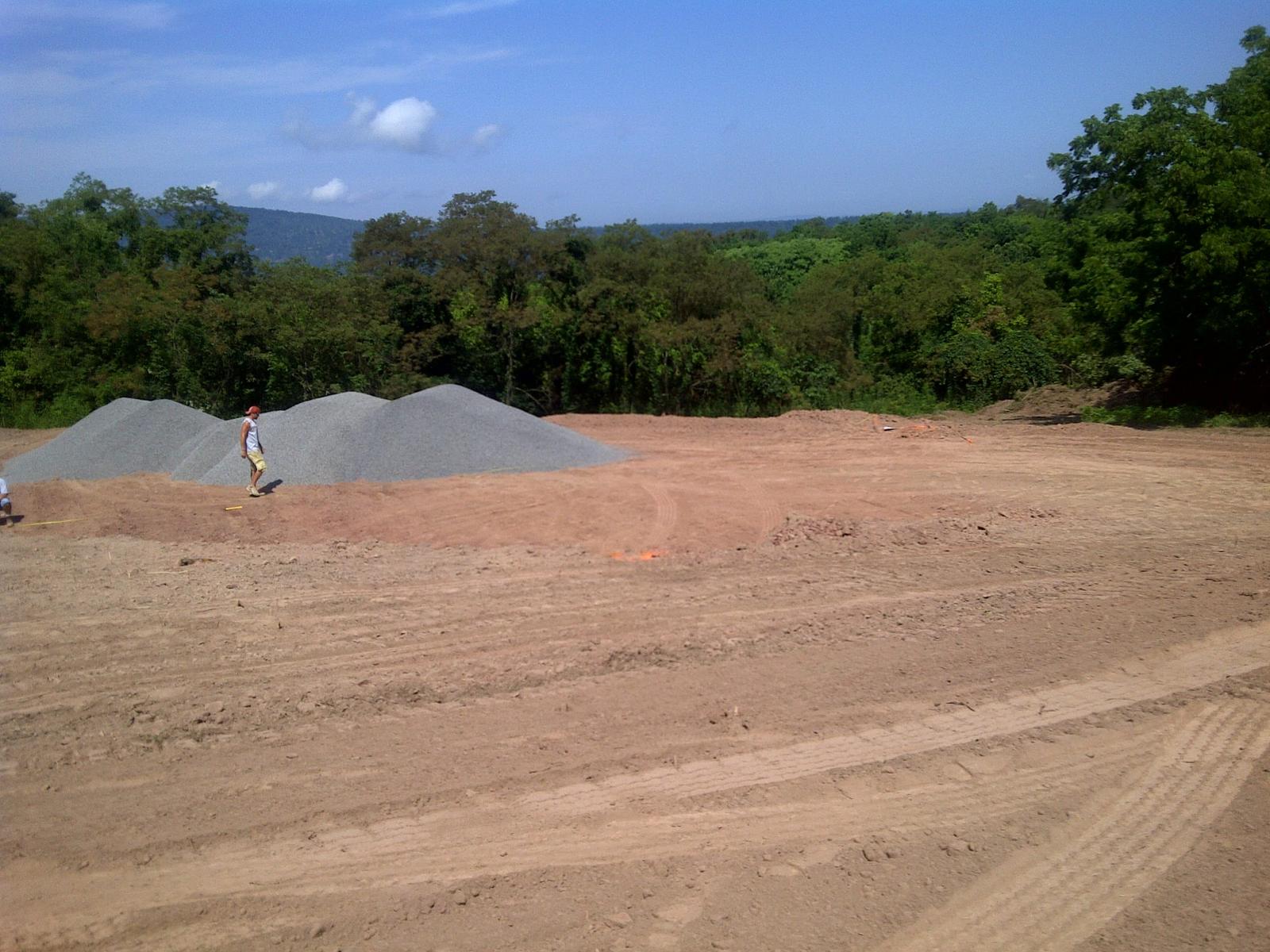 Gravel stockpiled on the graded pad with one person walking beside it, distant ridge above the trees