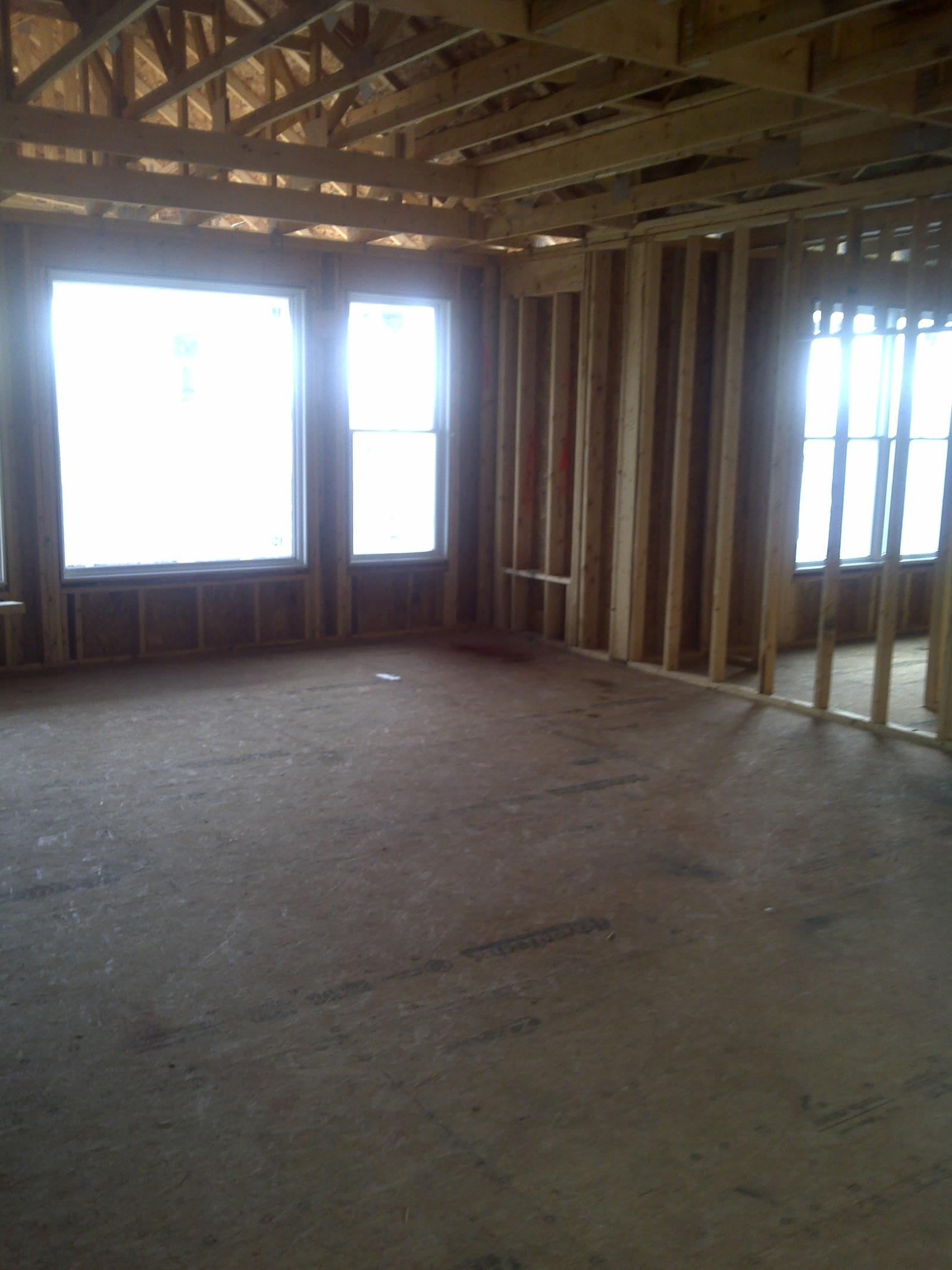 A framed room with three tall window openings letting in white daylight, bare subfloor underfoot