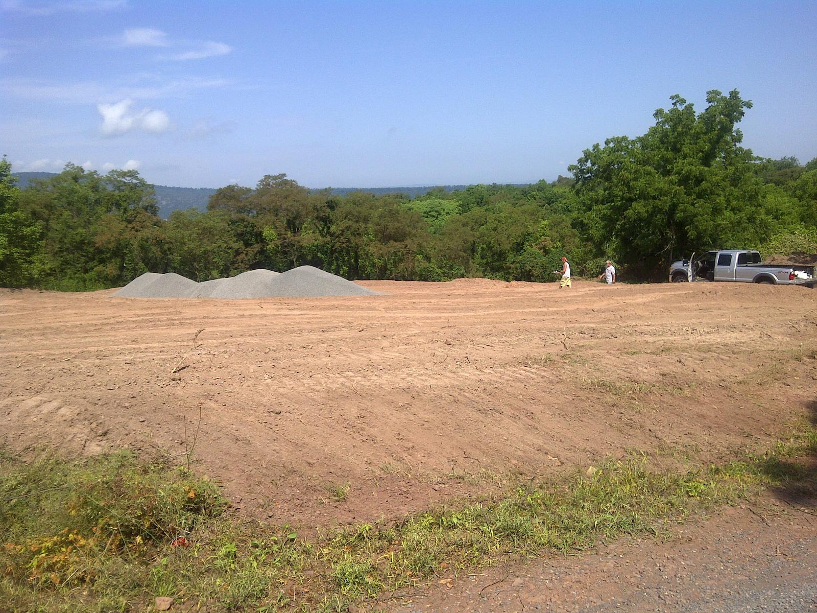 A graded building pad on a hilltop, gravel stockpiled at one end and a pickup at the other, cleared treeline and hazy ridges beyond