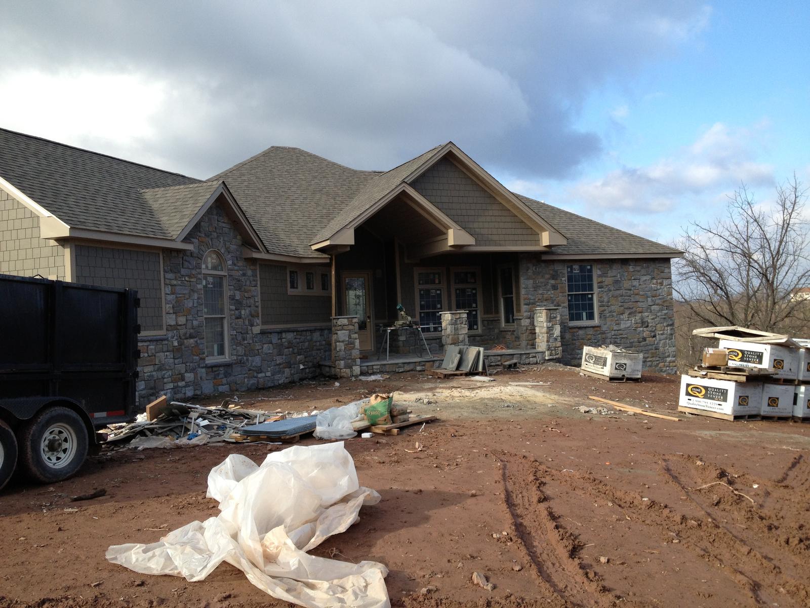 The house from the yard with stone up to the sills and roofing on, pallets and a dumpster standing in the mud