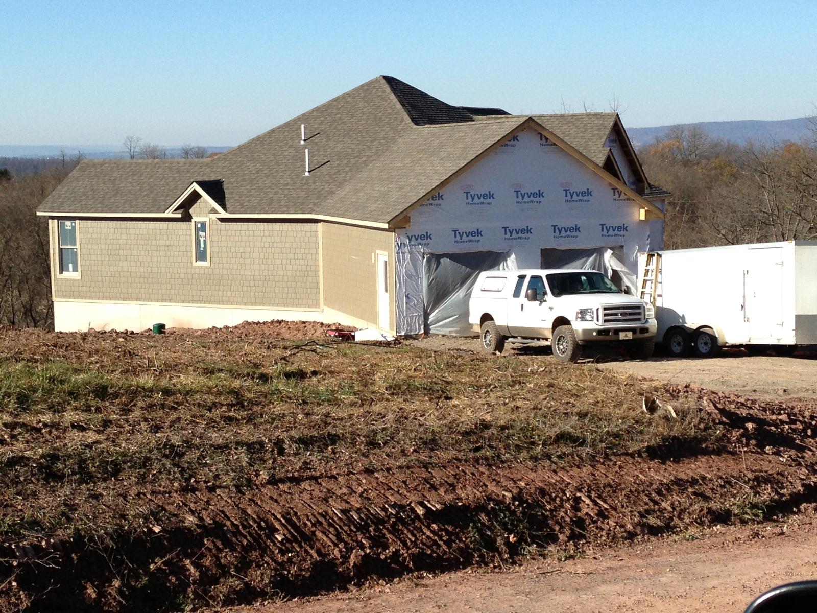The garage end under housewrap with a truck and trailer parked at the opening, fresh earth graded around