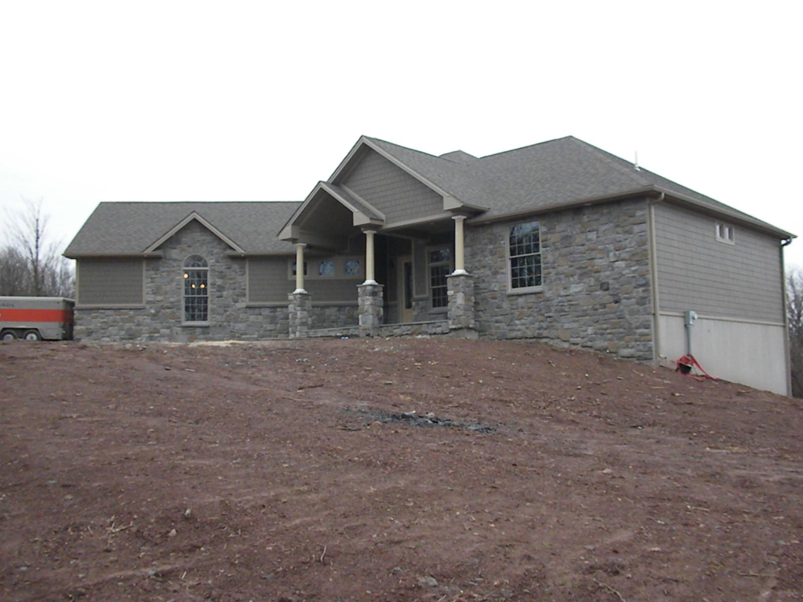 The house along its length from below, stone and shingle walls and a low-pitched roof, raw graded bank in front