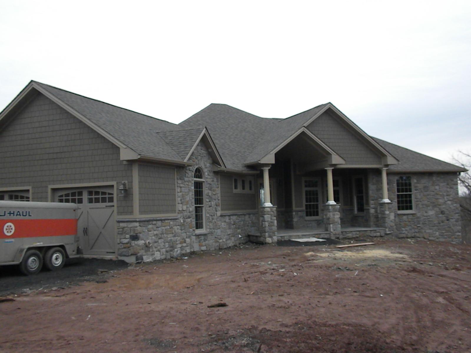 The house from the drive with its garage wing at the left, gray shingle and stone under a new roof, bare earth all around