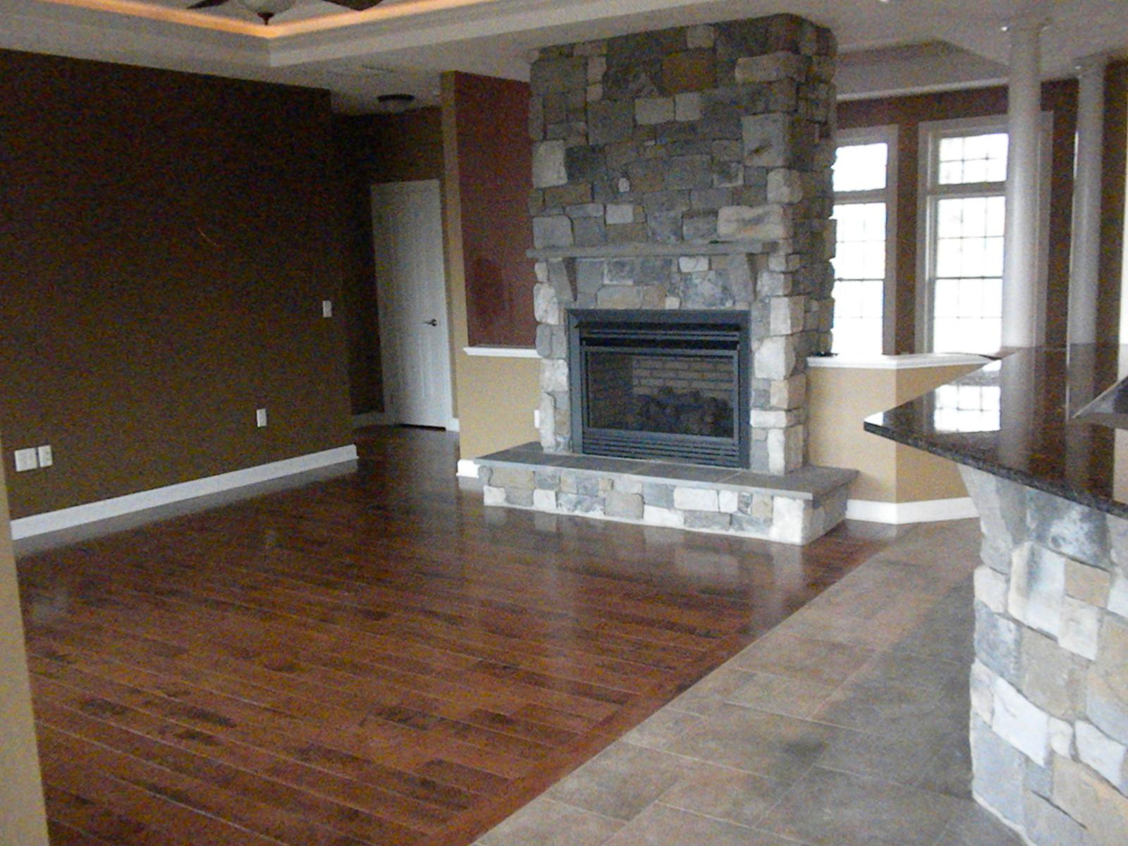 The great room from the entry, stone fireplace centered on the far wall between windows, wood floor giving way to tile