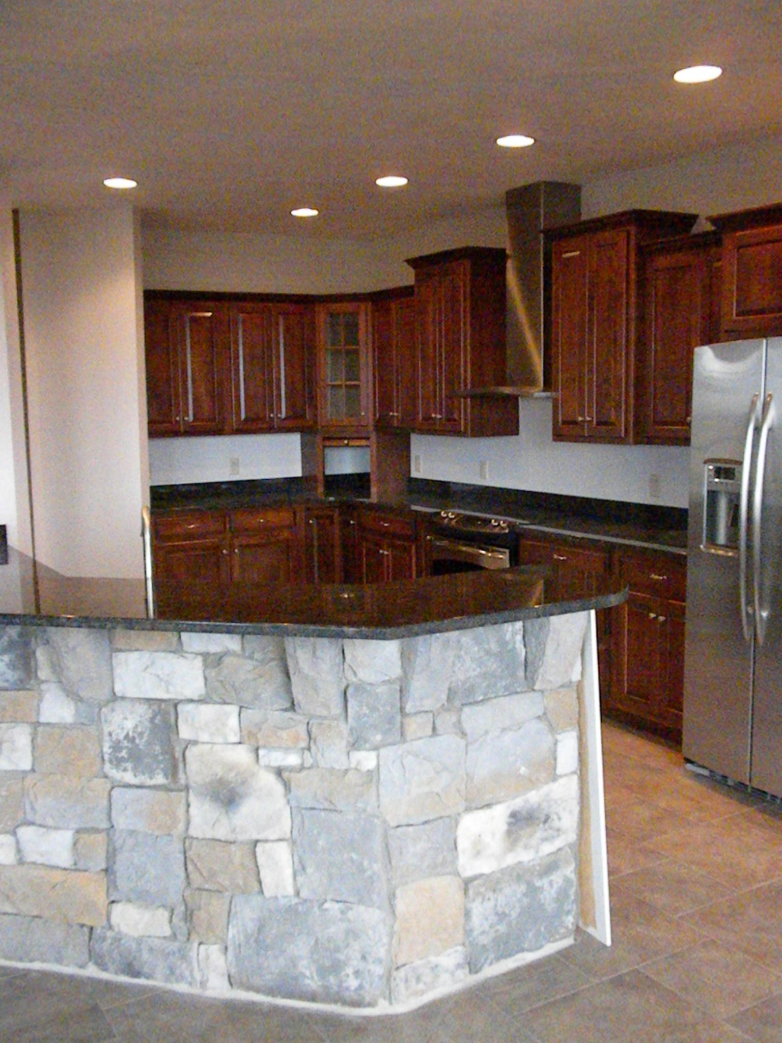 A kitchen with dark cabinets and a stone-faced island, wall oven and stainless refrigerator under recessed lights