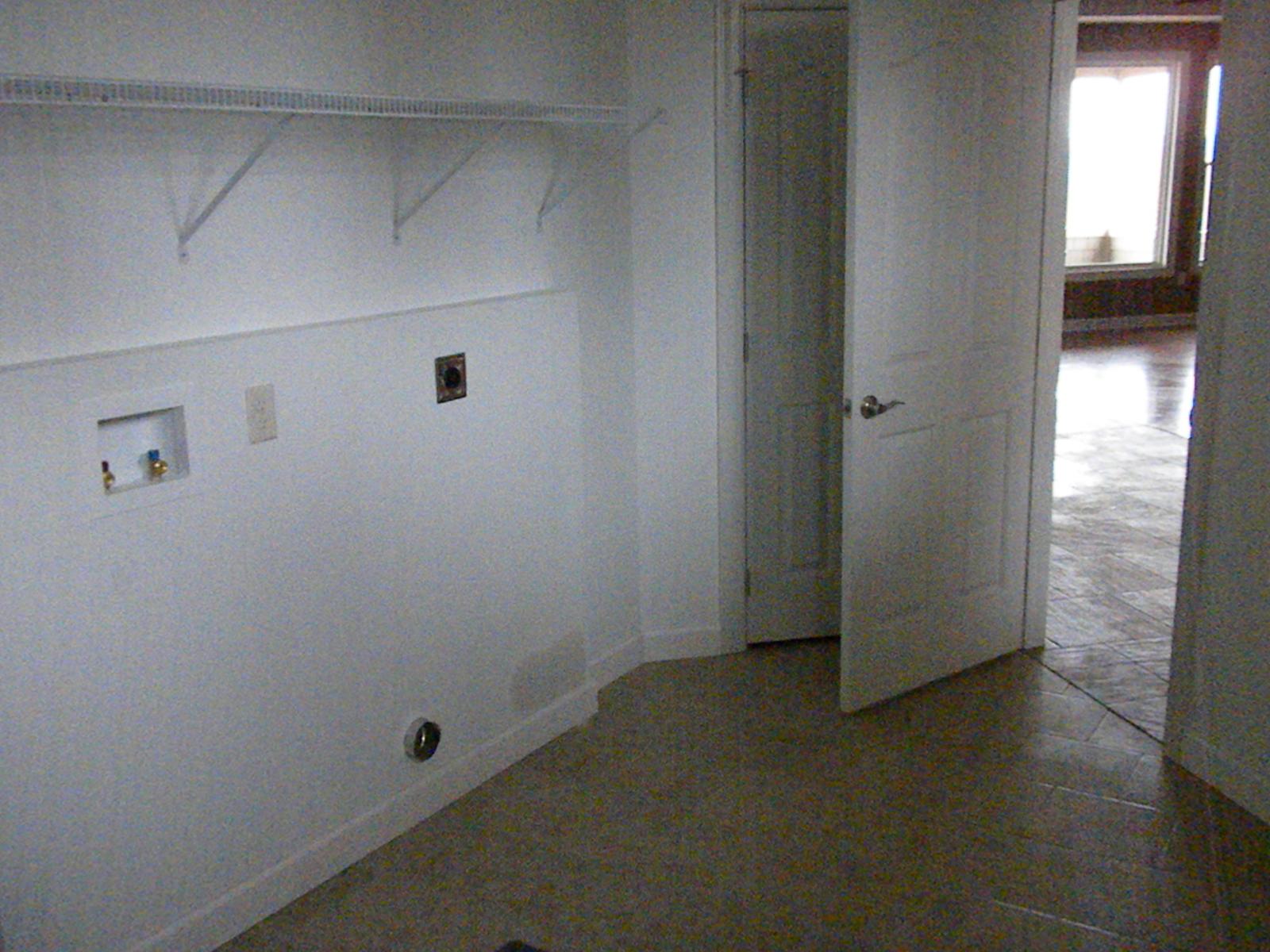 A laundry alcove with plumbing and outlets set in the wall, a shelf bracket above and a door standing open to a hall
