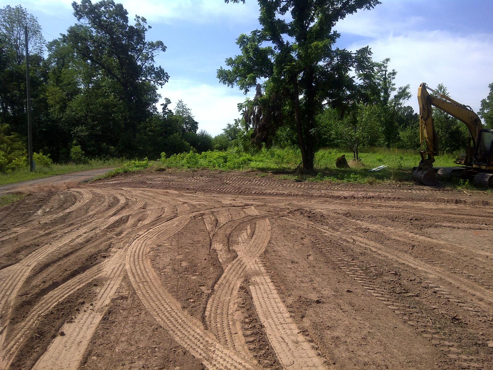Tire-rutted earth in the foreground with one large hardwood left standing, an excavator parked beyond it