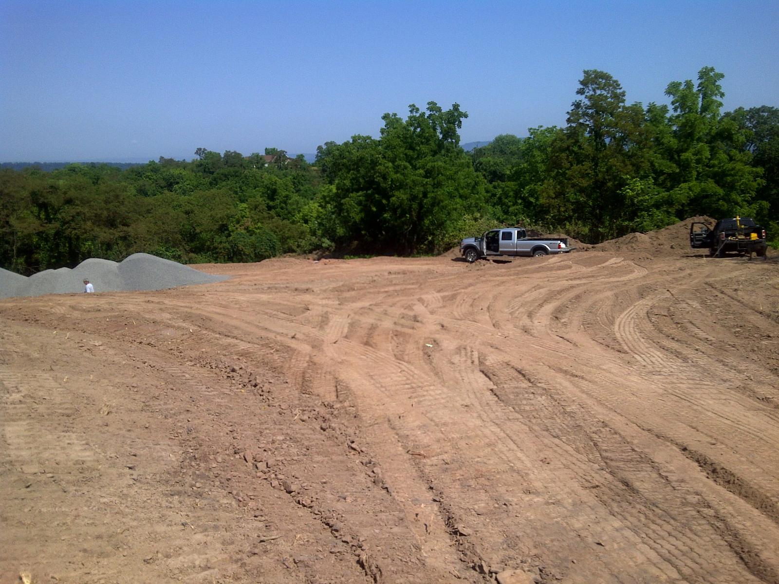 The graded pad from one corner, gravel stockpiles along the left and two vehicles parked at the treeline