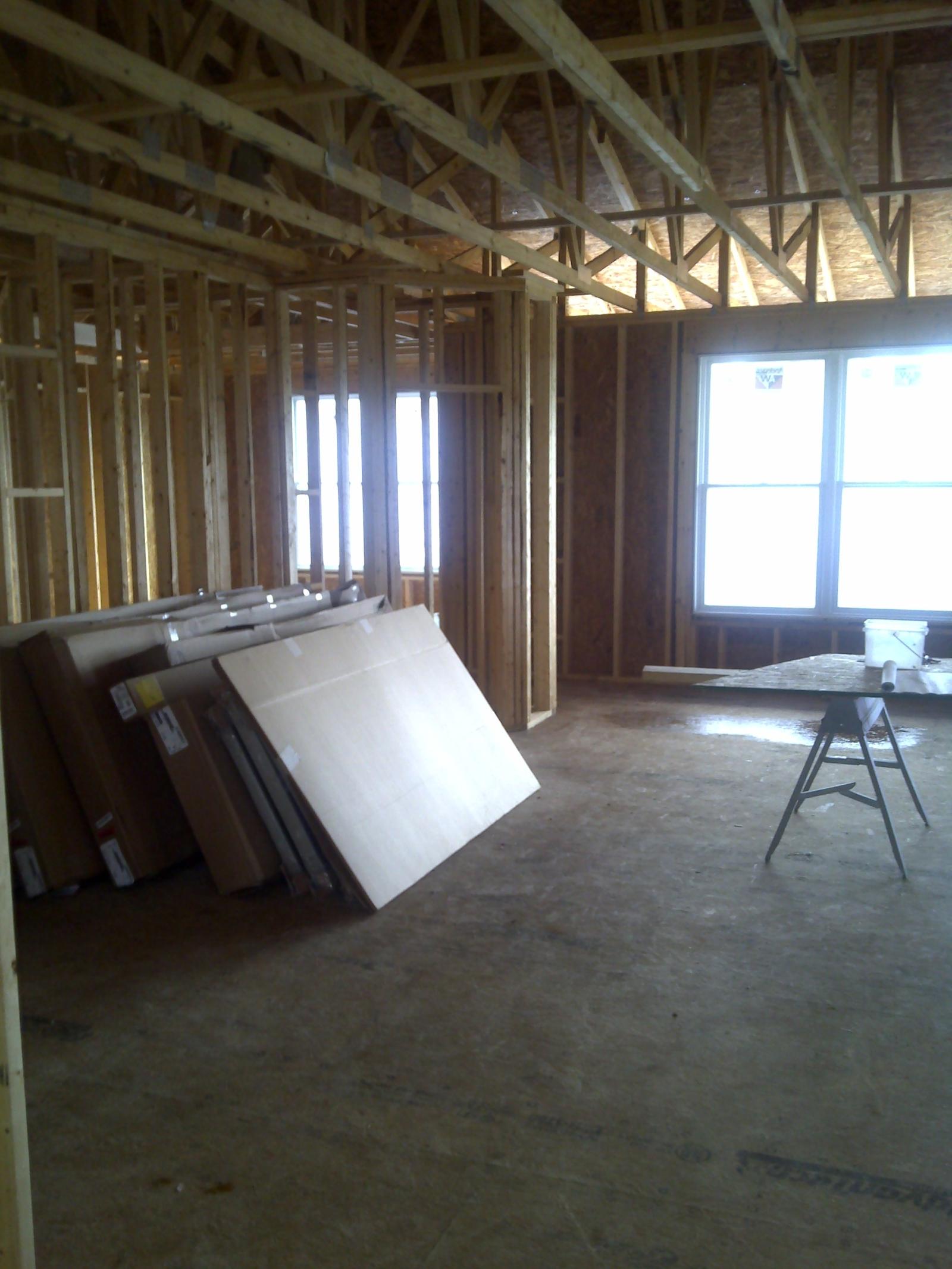 The interior framed and roofed, sheet goods stacked against a stud wall and a sawhorse standing in the open floor