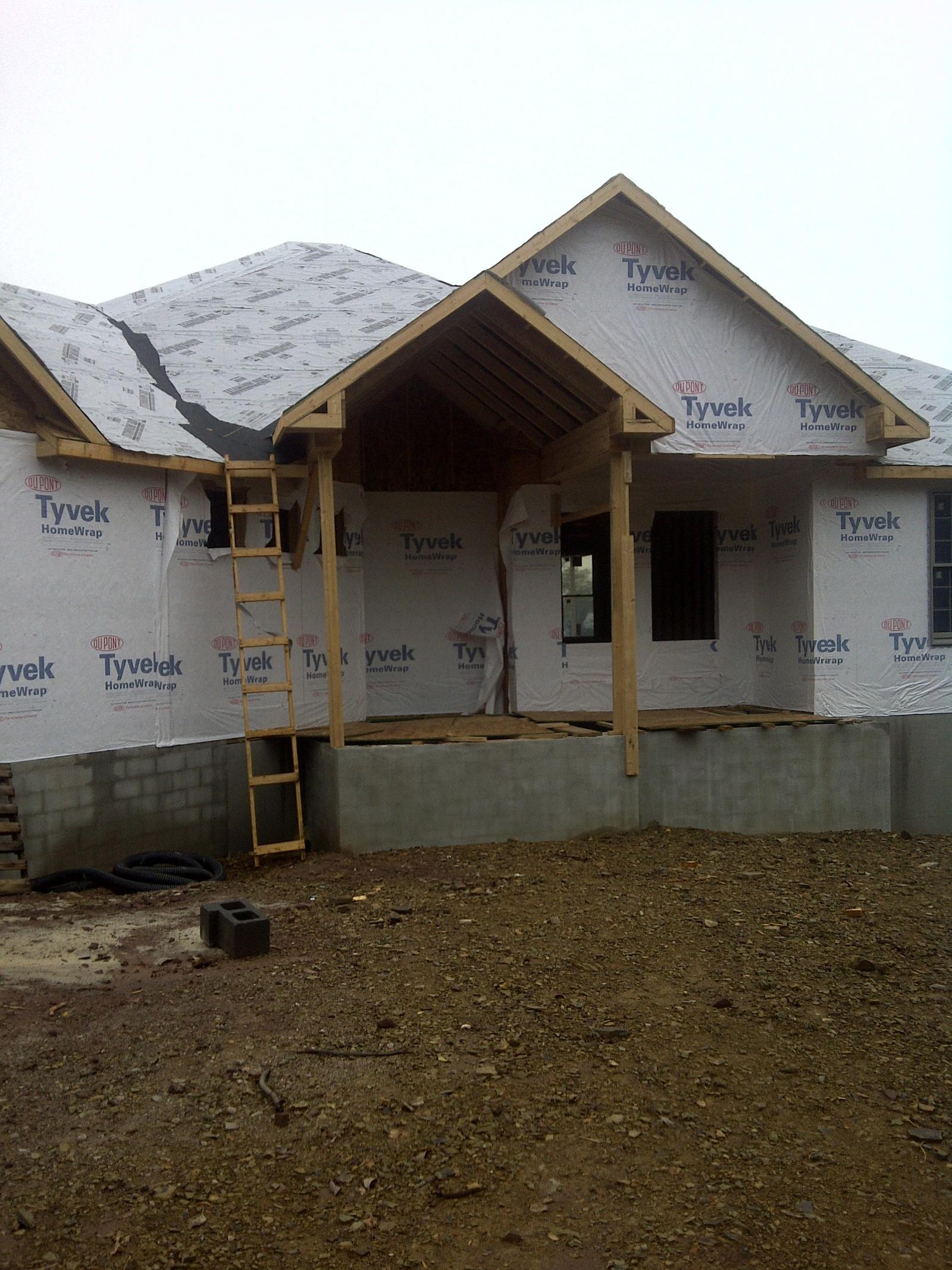 The house under housewrap with its porch gable framed, an extension ladder against the wall and block foundation below