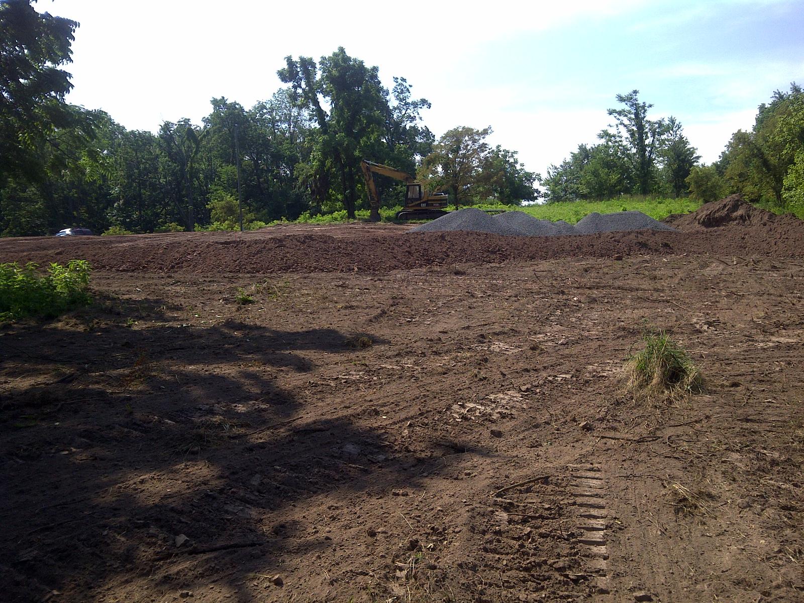 Freshly graded dark earth scored with track marks, an excavator working at the far edge against the trees