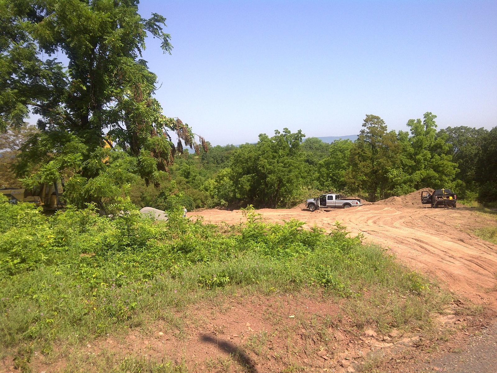 A new dirt access road cut through green scrub, a pickup and machinery parked along it, the valley opening on the right