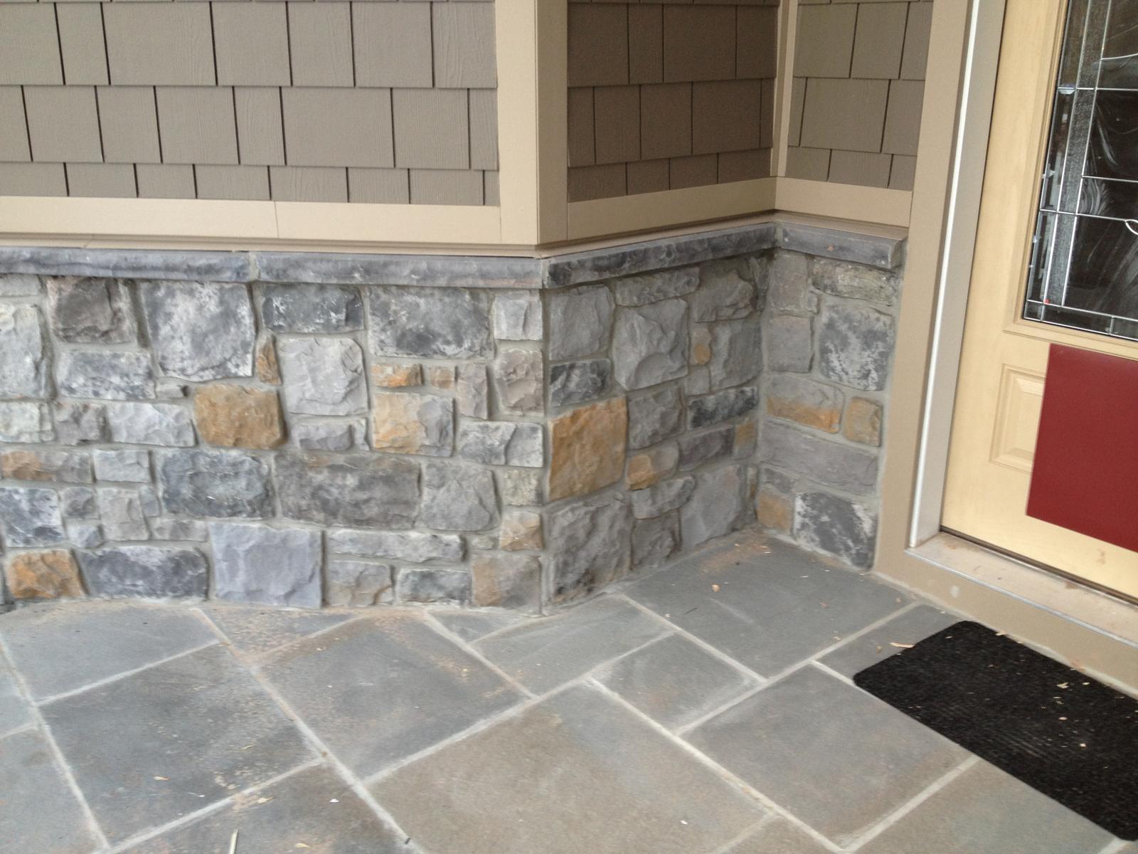 Flagstone paving meeting the stone wall at the porch corner, a red house-wrap patch beside the door