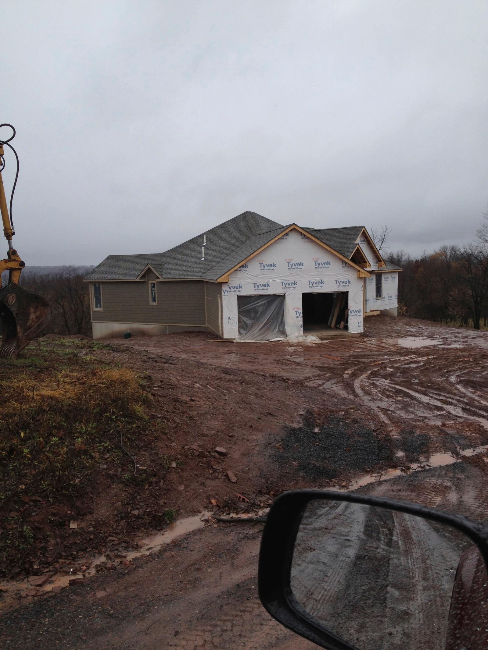 The house from the drive under housewrap, garage doors open, wet rutted ground in front